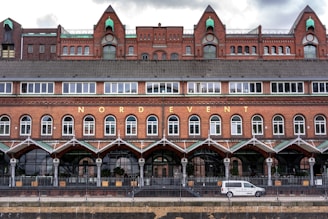 A large brick building with multiple levels and arched windows is seen with the word 'NORDEVENT' displayed prominently. The building features intricate architectural details including a dark tiled roof and decorative pillars. In front of the building, there is a white van parked, and the area appears to be fenced.