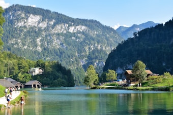 A serene lakeside landscape with lush green trees and towering mountains in the background. Several small wooden cabins are nestled among the greenery, and people are leisurely walking along the edge of the clear water. The sky is clear and blue, enhancing the tranquil and picturesque setting.