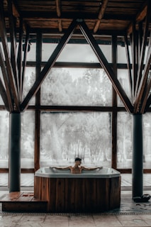 A person is relaxing in a hot tub situated indoors with large windows. The room features wooden beams and pillars, with an outdoors view of trees through the windows.