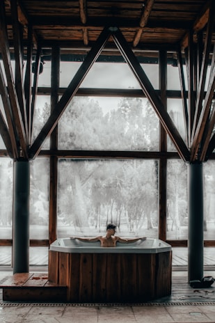 A person is relaxing in a hot tub situated indoors with large windows. The room features wooden beams and pillars, with an outdoors view of trees through the windows.
