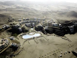 An aerial view of a desert resort complex featuring modern buildings arranged in clusters along winding roads. The center of the development includes two large swimming pools with curved shapes and landscaped areas. The surrounding environment consists of arid, mountainous terrain with sparse vegetation.