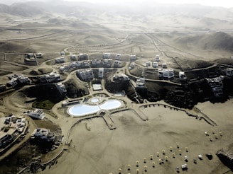 An aerial view of a desert resort complex featuring modern buildings arranged in clusters along winding roads. The center of the development includes two large swimming pools with curved shapes and landscaped areas. The surrounding environment consists of arid, mountainous terrain with sparse vegetation.