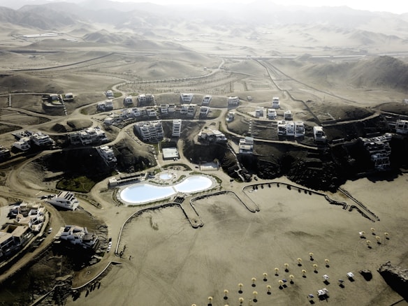 An aerial view of a desert resort complex featuring modern buildings arranged in clusters along winding roads. The center of the development includes two large swimming pools with curved shapes and landscaped areas. The surrounding environment consists of arid, mountainous terrain with sparse vegetation.