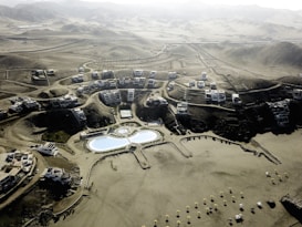 An aerial view of a desert resort complex featuring modern buildings arranged in clusters along winding roads. The center of the development includes two large swimming pools with curved shapes and landscaped areas. The surrounding environment consists of arid, mountainous terrain with sparse vegetation.