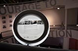 A circular frame with information and images displayed on a screen in a modern interior space. The background includes partial text from a large wall installation and black-and-white photographs. Soft lighting creates a contemporary and sophisticated environment.