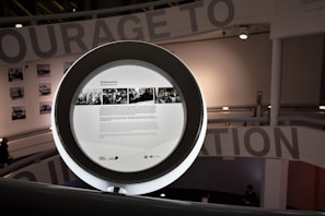 A circular frame with information and images displayed on a screen in a modern interior space. The background includes partial text from a large wall installation and black-and-white photographs. Soft lighting creates a contemporary and sophisticated environment.