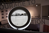 A circular frame with information and images displayed on a screen in a modern interior space. The background includes partial text from a large wall installation and black-and-white photographs. Soft lighting creates a contemporary and sophisticated environment.