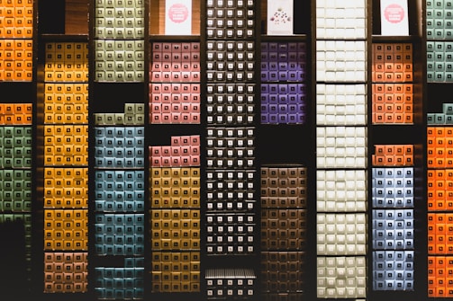 Various colorful stacked boxes are organized neatly on dark-colored shelves, creating a visually structured pattern. Each section of the shelves holds boxes of different colors, including greens, yellows, blues, purples, and oranges, contributing to a vibrant and orderly display.