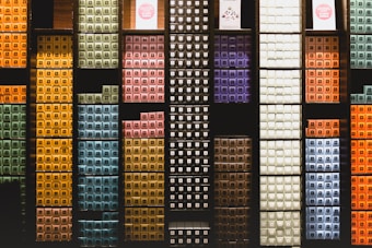 Various colorful stacked boxes are organized neatly on dark-colored shelves, creating a visually structured pattern. Each section of the shelves holds boxes of different colors, including greens, yellows, blues, purples, and oranges, contributing to a vibrant and orderly display.