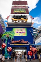 A vibrant and colorful entrance to an amusement attraction displaying a large sign labeled 'SlotZilla'. The entrance is adorned with imagery of oversized dice, palm trees, and a pink flamingo, all set against a bright blue backdrop. People are walking towards the entrance beneath the neon 'Fremont Street Experience' sign, creating a lively atmosphere.