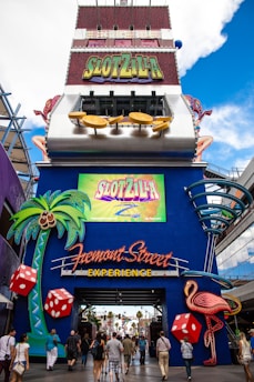 A vibrant and colorful entrance to an amusement attraction displaying a large sign labeled 'SlotZilla'. The entrance is adorned with imagery of oversized dice, palm trees, and a pink flamingo, all set against a bright blue backdrop. People are walking towards the entrance beneath the neon 'Fremont Street Experience' sign, creating a lively atmosphere.