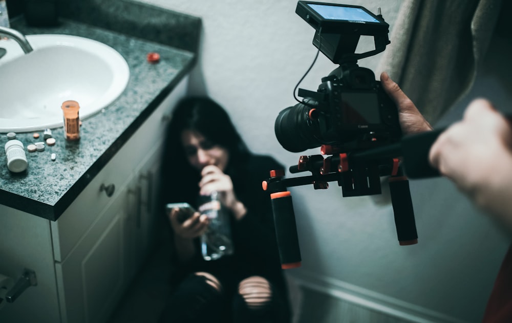 person taking video of woman sitting on wall in front of sink with cabinet