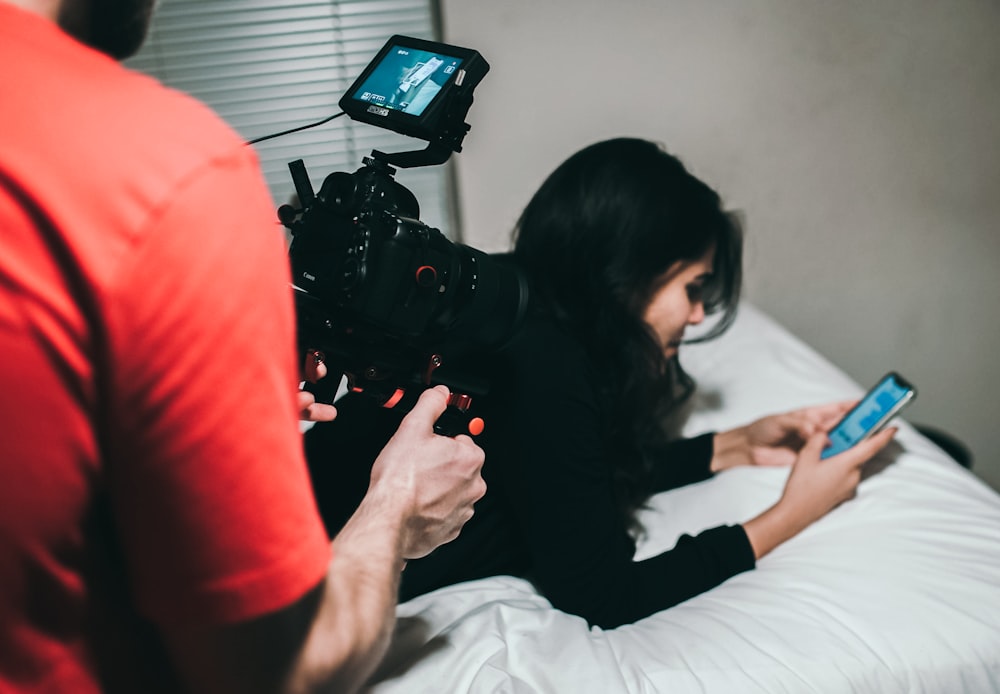 Mujer sosteniendo un teléfono inteligente mientras está acostada en la cama cerca de un hombre que sostiene una cámara profesional