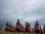 A group of people dressed in traditional, colorful attire with orange, black, and white patterns are gathered outdoors under a cloudy sky. They appear to be engaged in a ceremonial or festive activity, possibly dancing or marching, accompanied by musical instruments.
