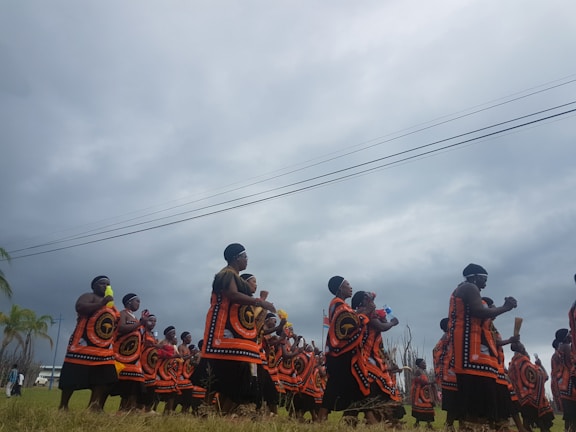 A group of people dressed in traditional, colorful attire with orange, black, and white patterns are gathered outdoors under a cloudy sky. They appear to be engaged in a ceremonial or festive activity, possibly dancing or marching, accompanied by musical instruments.