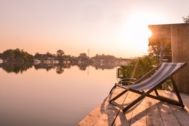 A serene lakeside setting during sunset with calm reflective waters and distant trees. A wooden deck features a striped deck chair facing the water, and the warm glow of the sun illuminates the scene, enhancing the tranquility.