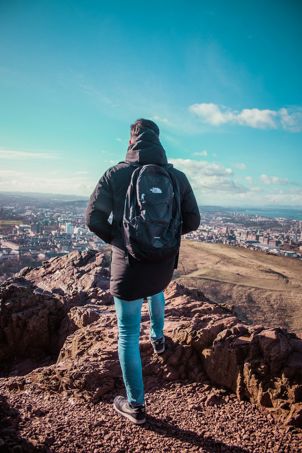 Persona in giacca con cappuccio e blue jeans che osserva la città sulla montagna rocciosa