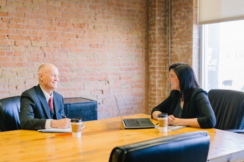 Two people are engaged in a conversation at a wooden conference table in a room with exposed brick walls. One is an older man in a suit and tie, holding a notebook, and the other is a woman with long dark hair, using a laptop. Both have coffee cups in front of them and are smiling.
