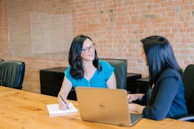 Duas mulheres conversando em uma mesa com um computador e um bloco de anotação