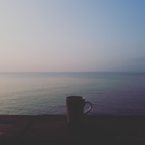 A tranquil scene featuring a calm sea with a soft gradient sky during either sunrise or sunset. A solitary mug is placed on a wooden surface in the foreground, enhancing the peaceful ambiance.