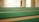 A spacious area with rows of green carpets laid out on a large floor, illuminated by natural light coming through windows. The walls are light-colored, contributing to a serene and open atmosphere.