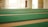 A spacious area with rows of green carpets laid out on a large floor, illuminated by natural light coming through windows. The walls are light-colored, contributing to a serene and open atmosphere.