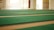 A spacious area with rows of green carpets laid out on a large floor, illuminated by natural light coming through windows. The walls are light-colored, contributing to a serene and open atmosphere.