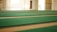 A spacious area with rows of green carpets laid out on a large floor, illuminated by natural light coming through windows. The walls are light-colored, contributing to a serene and open atmosphere.