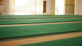 A spacious area with rows of green carpets laid out on a large floor, illuminated by natural light coming through windows. The walls are light-colored, contributing to a serene and open atmosphere.