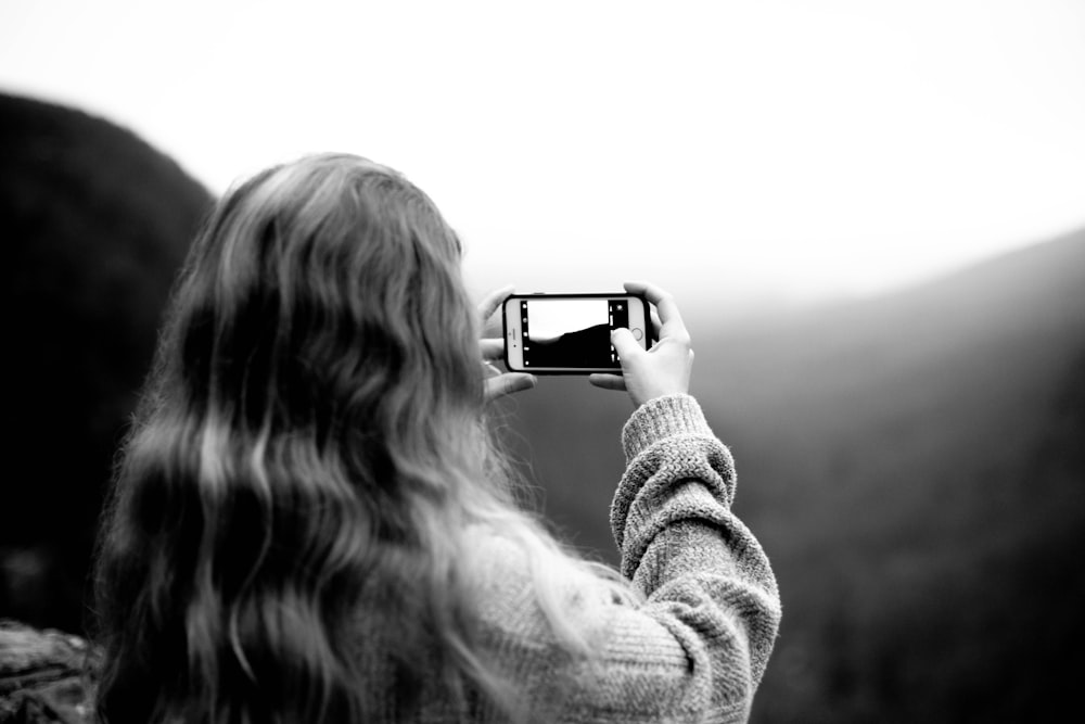 grayscale photography of woman wearing sweater using smartphone taking picture