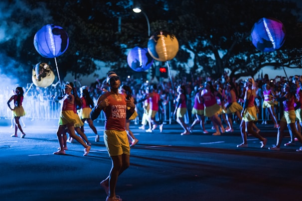 A vibrant parade scene at night with a group of people wearing matching outfits, featuring red tops and yellow skirts. They appear to be holding large, globe-shaped lanterns. The street is illuminated by colorful lighting and there are crowds watching from behind barricades.