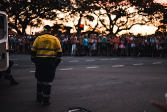 A traffic emergency patrol officer stands on a street facing a large, gathered crowd behind safety barriers. The crowd appears to be at an event or parade during sunset, with a van nearby and silhouettes of large trees in the background.