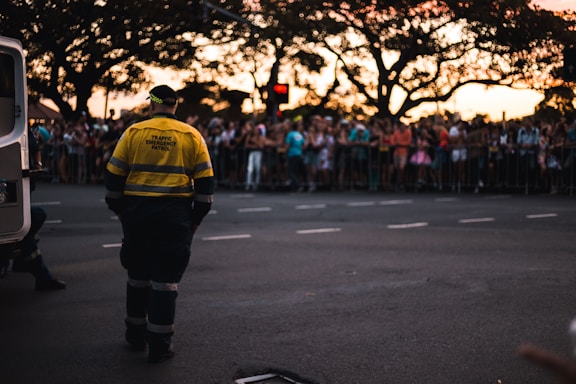 A traffic emergency patrol officer stands on a street facing a large, gathered crowd behind safety barriers. The crowd appears to be at an event or parade during sunset, with a van nearby and silhouettes of large trees in the background.