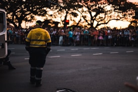 A traffic emergency patrol officer stands on a street facing a large, gathered crowd behind safety barriers. The crowd appears to be at an event or parade during sunset, with a van nearby and silhouettes of large trees in the background.