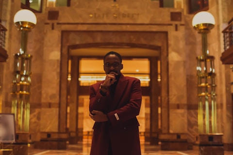 A person wearing a dark red coat stands in an elegant, warmly lit interior space with marble walls and shiny golden columns. The atmosphere is luxurious and sophisticated, with a mixture of modern and classic architectural elements.