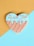 A heart-shaped cookie is decorated with a light blue icing on the top half, adorned with colorful sprinkles. The bottom half of the cookie has a waffle pattern in light brown. The background features a diagonal split between mustard yellow and pastel blue colors.