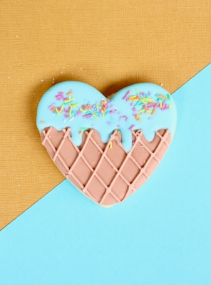 A heart-shaped cookie is decorated with a light blue icing on the top half, adorned with colorful sprinkles. The bottom half of the cookie has a waffle pattern in light brown. The background features a diagonal split between mustard yellow and pastel blue colors.