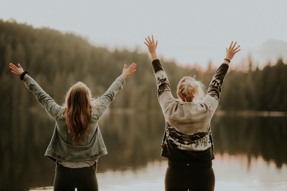 two women hands up standing beside body of water