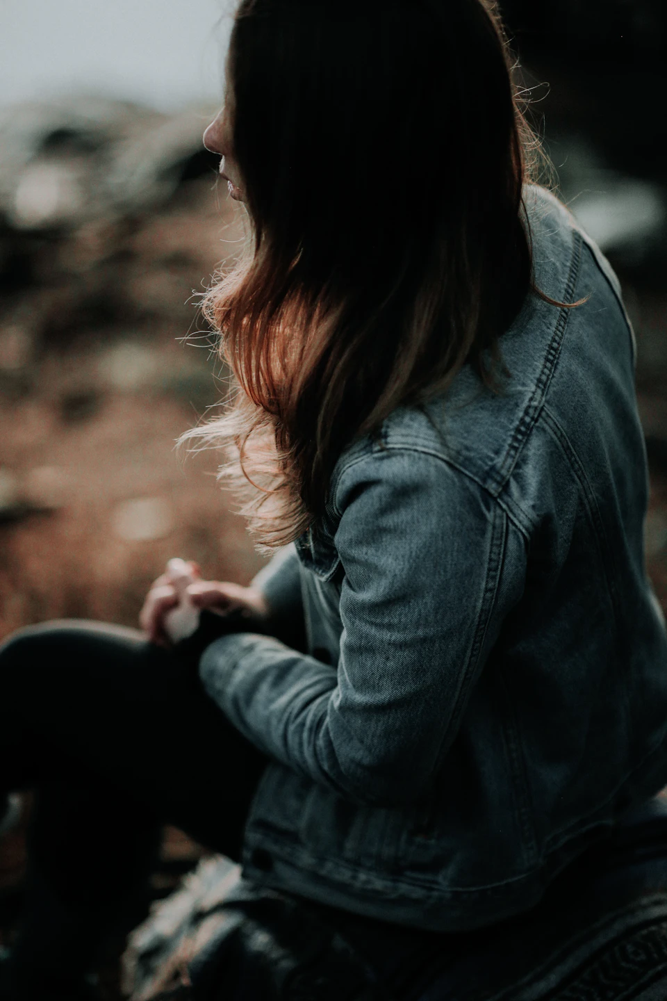 Young woman sits on a rock with denim jacket and black pants facing away