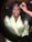 A woman in a light green robe is having makeup applied by another person using a brush. She is seated in front of a lit mirror, wearing dark curly hair and exhibits a calm expression.