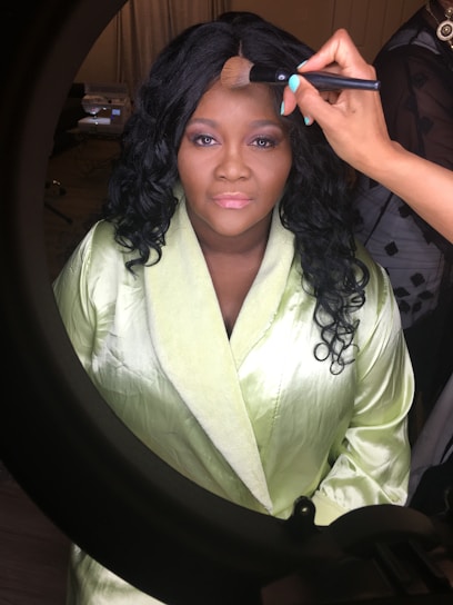 A woman in a light green robe is having makeup applied by another person using a brush. She is seated in front of a lit mirror, wearing dark curly hair and exhibits a calm expression.