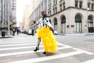 A person confidently walks across a city street wearing a bold outfit featuring a black and white polka dot top and a vibrant yellow tulle skirt. The leggings are patterned with black and yellow lettering, and the ensemble is completed with high-heeled shoes. Surrounding buildings are tall and architecturally detailed, with several pedestrians and parked cars visible in the background.