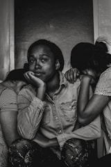 A black and white photograph depicting three individuals. The central figure, with a contemplative expression, rests their chin on their hand while seated. Two other people lean on either side, with their heads tucked in, creating a sense of intimacy and support. The mood is introspective and somber.