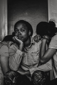 A black and white photograph depicting three individuals. The central figure, with a contemplative expression, rests their chin on their hand while seated. Two other people lean on either side, with their heads tucked in, creating a sense of intimacy and support. The mood is introspective and somber.