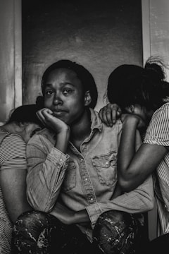 A black and white photograph depicting three individuals. The central figure, with a contemplative expression, rests their chin on their hand while seated. Two other people lean on either side, with their heads tucked in, creating a sense of intimacy and support. The mood is introspective and somber.