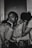 A black and white photograph depicting three individuals. The central figure, with a contemplative expression, rests their chin on their hand while seated. Two other people lean on either side, with their heads tucked in, creating a sense of intimacy and support. The mood is introspective and somber.