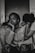 A black and white photograph depicting three individuals. The central figure, with a contemplative expression, rests their chin on their hand while seated. Two other people lean on either side, with their heads tucked in, creating a sense of intimacy and support. The mood is introspective and somber.