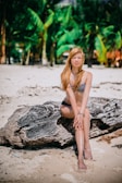 A woman in a bikini is sitting on a large piece of driftwood on a sandy beach. She has long blonde hair and is posing with her hands clasped over her knees. The background features lush green palm trees, indicating a tropical setting.