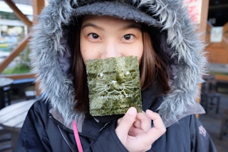 A person wearing a hooded coat holds a piece of seaweed in front of their face. The seaweed features a cute illustration of a cat face and the text 'Asahiyama Zoo' printed on it. Their eyes are visible above the seaweed, and there is a blurred outdoor setting in the background with tables and chairs.