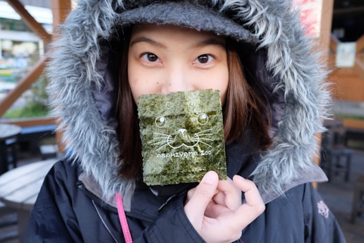 A person wearing a hooded coat holds a piece of seaweed in front of their face. The seaweed features a cute illustration of a cat face and the text 'Asahiyama Zoo' printed on it. Their eyes are visible above the seaweed, and there is a blurred outdoor setting in the background with tables and chairs.